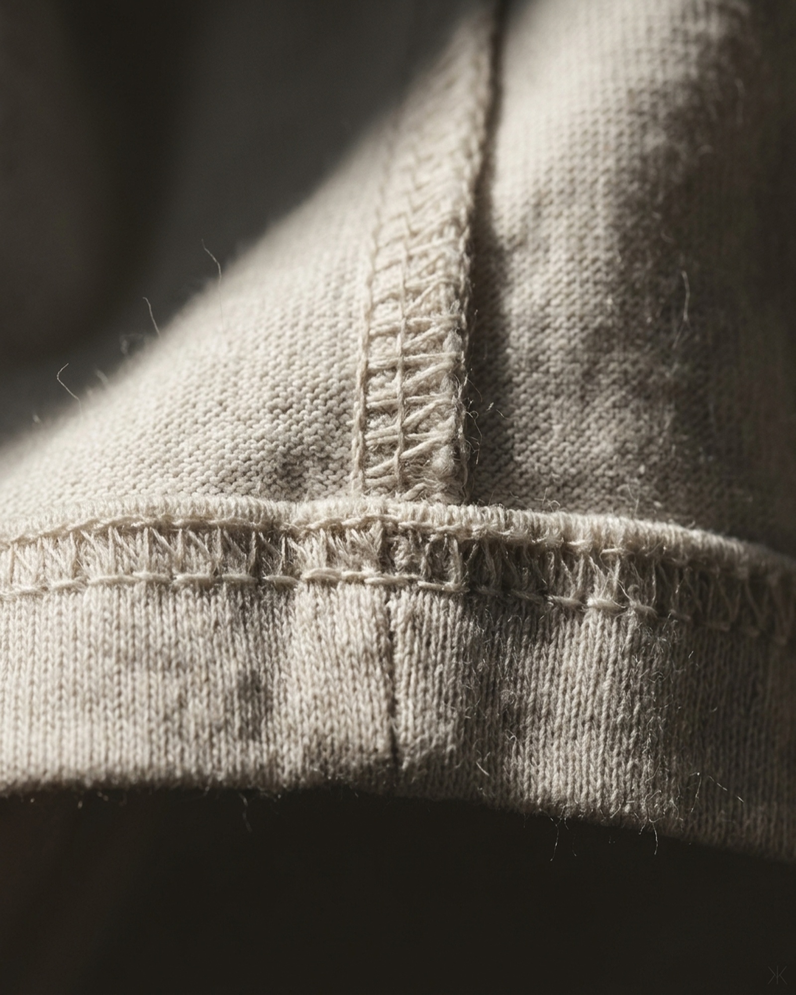 Extreme macro of a cotton jersey t-shirt hem and overlocked side seam — visible knit loops and thread tension, raw natural cotton color, one hard directional shaft of light grazing the surface, deep falloff to shadow. No tags, no logos, no text.